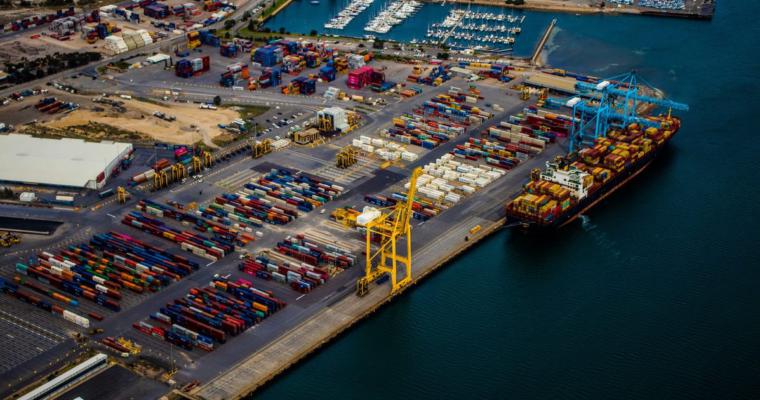 Luftaufnahme eines Containerterminals, auf dem Tausende von Containern auf dem Gelände gestapelt sind, ein einzelnes Schiff am Kai liegt und beladen wird und ein Kran am Kai stillsteht